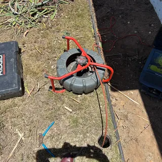 A RIDGID SeeSnake camera reel feeding push cable into an open cleanout in a lawn, with the locator case alongside and the monitor showing the inside of the line