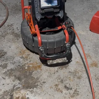 A camera reel with the monitor mounted on it, orange push cable running out toward a broken-out opening in the concrete floor, the screen showing the inside of the pipe