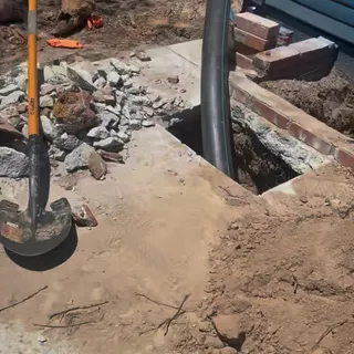 An open excavation beside a house foundation, concrete broken out and stacked brick shoring the edge, with the sewer line exposed at the bottom