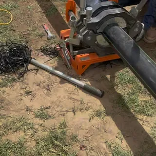 A butt-fusion machine clamped onto two lengths of black HDPE pipe on a lawn, with the shavings from facing the pipe ends coiled on the ground beside it