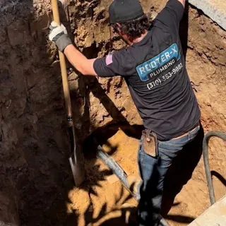 A Rooter-X crew member standing in a shored excavation, digging by hand with a long shovel down toward the line, hydraulic hoses running over the edge above